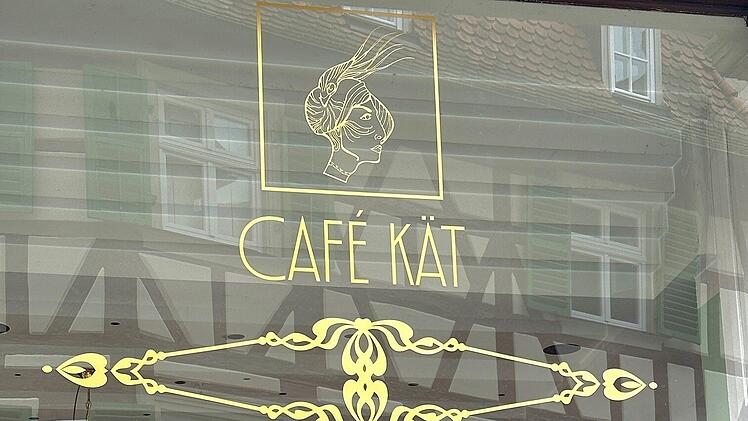 Caf&eacute; K&auml;t in Bamberg: Goldene Verzierung auf Fensterscheibe, stilvolles Logo mit Frauensilhouette. Dahinter sieht man Fachwerk und einen Kronleuchter.