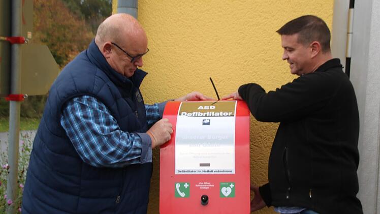 Ortsreferent Klaus Schebler (links) und der der Vorsitzende des Feuerwehrvereins, Heiko Müller, bringen den Defibrillator am Feuerwehrgerätehaus an. Die Gerätehäuser werden jetzt auch ans schnelle Netz angeschlossen. Thomas Malz