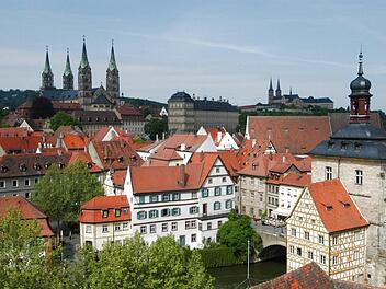Bamberg mit seinen markanten roten Dächern und historischen Bauwerken, darunter das Alte Rathaus auf der Brücke. Die Skyline ist geprägt von Kirchtürmen und üppigem Grün.