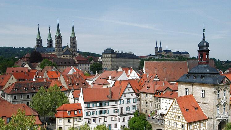 Bamberg mit seinen markanten roten Dächern und historischen Bauwerken, darunter das Alte Rathaus auf der Brücke. Die Skyline ist geprägt von Kirchtürmen und üppigem Grün.