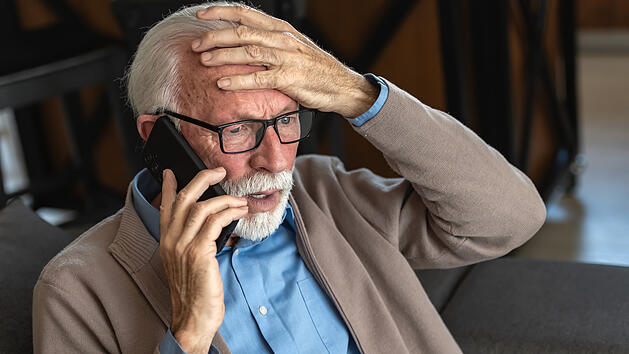 An elderly man with glasses and a beard, wearing a blue shirt and beige cardigan, looks distressed while talking on the phone, holding his head in concern