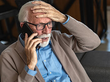 An elderly man with glasses and a beard, wearing a blue shirt and beige cardigan, looks distressed while talking on the phone, holding his head in concern
