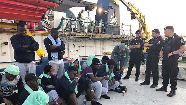 Migranten vom Rettungsschiff &laquo;Sea-Watch 3&raquo; sitzenbewacht von Polizisten auf dem Steg im Hafen der Insel Lampedusa. Das Schiff der deutschen Hilfsorganisation Sea-Watch ist nach ddem unerlaubten Anlegen im Hafen der italienischen Insel Lampedusa und der Festnahme der Kapit&auml;nin beschlagnahmt worden. Das best&auml;tigte ein Sea-Watch-Sprecher. Foto: Annalisa Camilli/AP/dpa