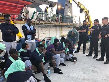 Migranten vom Rettungsschiff «Sea-Watch 3» sitzenbewacht von Polizisten auf dem Steg im Hafen der Insel Lampedusa. Das Schiff der deutschen Hilfsorganisation Sea-Watch ist nach ddem unerlaubten Anlegen im Hafen der italienischen Insel Lampedusa und der Festnahme der Kapitänin beschlagnahmt worden. Das bestätigte ein Sea-Watch-Sprecher. Foto: Annalisa Camilli/AP/dpa