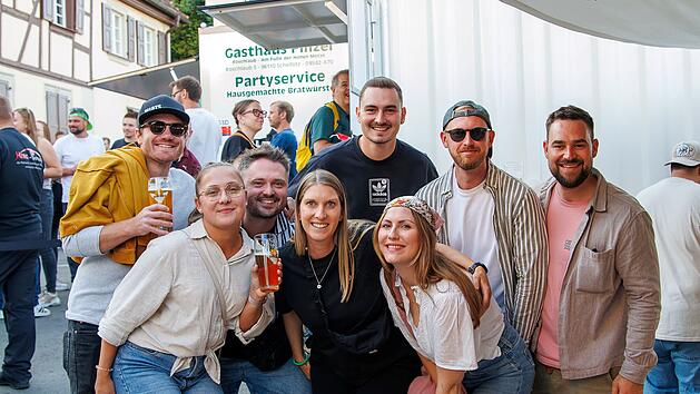 Fr&ouml;hliche Gruppe junger Menschen auf einem Volksfest. Sie halten Getr&auml;nke und posieren lachend vor einem Imbissstand. Im Hintergrund sind weitere Festivalbesucher zu sehen.
