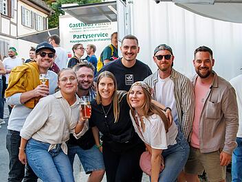 Fr&ouml;hliche Gruppe junger Menschen auf einem Volksfest. Sie halten Getr&auml;nke und posieren lachend vor einem Imbissstand. Im Hintergrund sind weitere Festivalbesucher zu sehen.