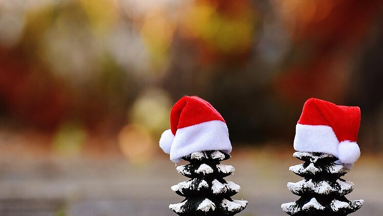 Zwei Tannenzapfen mit weihnachtlichen Mützen stehen auf einem Boden. Der Hintergrund ist unscharf und herbstlich gefärbt.