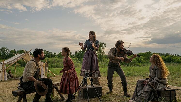 Fr&ouml;hliche Zeiten in der Pr&auml;rie: Schon in den B&uuml;chern griff Vater Charles Ingalls (Luke Bracey, zweiter von rechts), gerne zur Fiddle und erfreute damit seine T&ouml;chter Laura (Alice Halsey, zweite von links), Mary (Skywalker Hughes, Mitte), Ehefrau Caroline (Crosby Fitzgerald) und Familienfreund Mr. Edwards (Warren Christie).