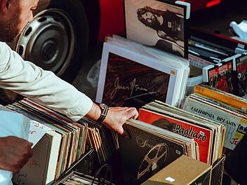 Person stöbert auf einem Flohmarkt durch Plattenkisten. Vintage-Schallplatten sind zu sehen, im Hintergrund ein Auto.