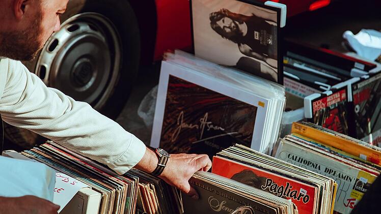Person stöbert auf einem Flohmarkt durch Plattenkisten. Vintage-Schallplatten sind zu sehen, im Hintergrund ein Auto.