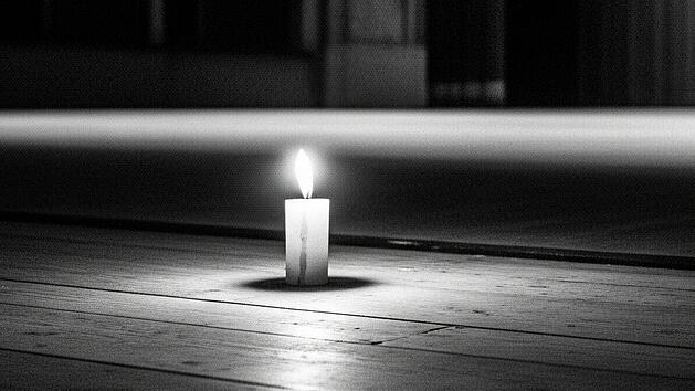 Auf der ansonsten leeren B&uuml;hne eines gro&szlig;en Theaters brennt eine Kerze. Das Bild ist ein Trauerbild in Schwarz-Wei&szlig;.