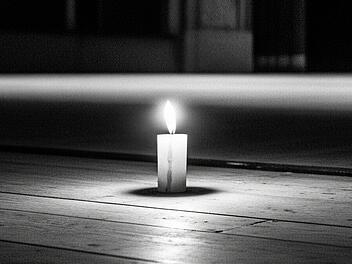 Auf der ansonsten leeren Bühne eines großen Theaters brennt eine Kerze. Das Bild ist ein Trauerbild in Schwarz-Weiß.