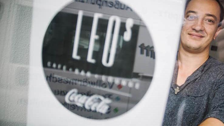Probehalber hält Oleg Knoll einen Design-Entwurf für das neue Logo an die Glastür des Lokals. In der Spiegelung ist der Transporter der Handwerker zu sehen, die gerade die letzten Arbeiten verrichten.  Foto: Steffens