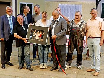 Vor dem Bühnenbild von "Widder Dahaam" präsentiert Martin Ritter (Zweiter von links) auf der Bühne eines seiner Szenenfotos der diesjährigen Theaterstücke. Mit dabei (von rechts) Vorsitzender Siegfried Küspert, Dieter Krause, Walter Richter, Werner Eberhardt (verdeckt), Silke Neukam-Ködel, Mathias Ebert, Gerd Kammerer, Bürgermeister Werner Diersch. Foto: Dieter Hübner