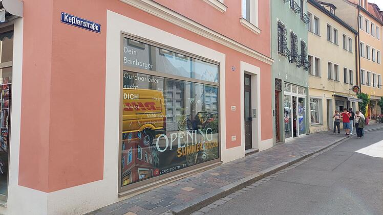 Ein neues Outdoor-Gesch&auml;ft er&ouml;ffnet im Sommer in der Ke&szlig;lerstra&szlig;e, Bamberg, in einem rosa Geb&auml;ude. Schaufensterwerbung ist sichtbar. Passanten sind auf der ruhigen Stra&szlig;e unterwegs.
