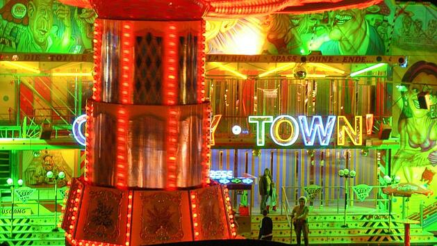 Ein hell erleuchteter Jahrmarkt mit bunten Lichtern und einer farbenfrohen Fassade. Ein Fahrgesch&auml;ft und der Schriftzug "Crazy Town" sind zu sehen. Ein Klassiker des Volksfestes.