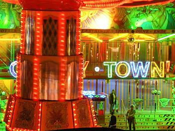 Ein hell erleuchteter Jahrmarkt mit bunten Lichtern und einer farbenfrohen Fassade. Ein Fahrgeschäft und der Schriftzug "Crazy Town" sind zu sehen. Ein Klassiker des Volksfestes.