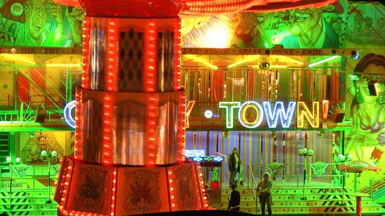 Ein hell erleuchteter Jahrmarkt mit bunten Lichtern und einer farbenfrohen Fassade. Ein Fahrgeschäft und der Schriftzug "Crazy Town" sind zu sehen. Ein Klassiker des Volksfestes.