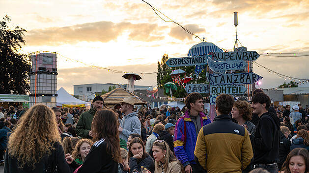 Das Bild zeigt ein lebhaftes Festival mit vielen Besuchern, Essenst&auml;nden und einer Au&szlig;enbar. Ein Schild weist auf verschiedene Attraktionen hin. Der Himmel ist teils bew&ouml;lkt beim Sonnenuntergang.