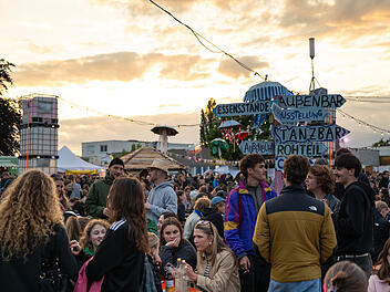 Das Bild zeigt ein lebhaftes Festival mit vielen Besuchern, Essenst&auml;nden und einer Au&szlig;enbar. Ein Schild weist auf verschiedene Attraktionen hin. Der Himmel ist teils bew&ouml;lkt beim Sonnenuntergang.