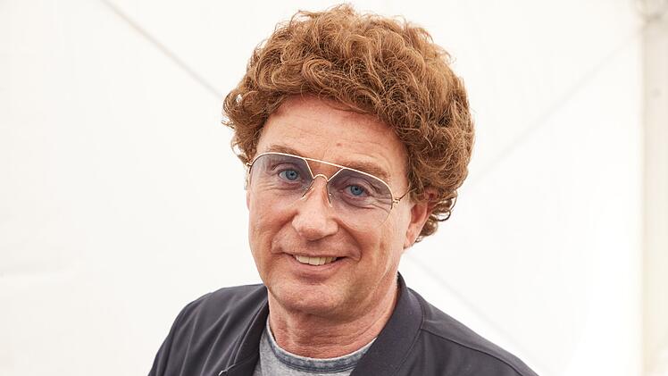 Eine Person mit lockigem Haar und Brille blickt lächelnd in die Kamera. Sie trägt eine dunkle Jacke und ein T-Shirt. Hintergrund ist schlicht und hell.