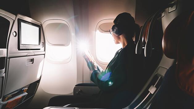 Female tourist in headphones listening audio book and looking in airplane window enjoying flight. Young 25 years old woman with cellular phone in hands sitting on seat during flight in first class Weibliche Touristin mit Kopfh&ouml;rern h&ouml;rt ein H&ouml;rbuch und schaut aus dem Flugzeugfenster, um den Flug zu genie&szlig;en. Junge 25-j&auml;hrige Frau mit Handy in der Hand sitzt w&auml;hrend des Fluges in der First Class auf ihrem Sitzplatz.