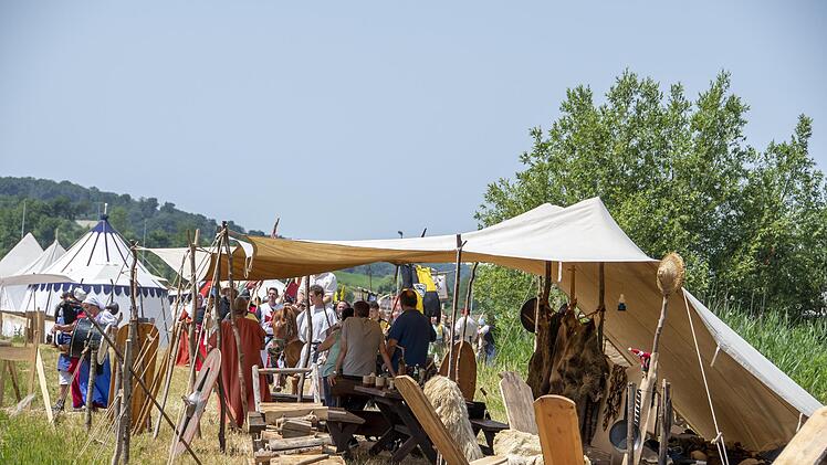 Menschen in mittelalterlichen Gewändern lagern in Zelten. Es gibt Holzmöbel und historische Ausrüstungsgegenstände unter blauem Himmel, umgeben von Wiesen.