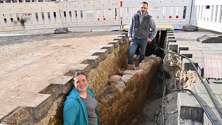 Zwei Personen stehen in einem Graben auf einer Baustelle, umgeben von Absperrungen. Der Graben zeigt Erdschichten und freigelegte, alte Steine.