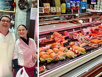 Zwei Personen in Metzgerei vor Fleischtheke. Schilder mit Preisen und Angeboten, darunter Sandwiches und Würste. Weihnachtliche Dekoration im Hintergrund.