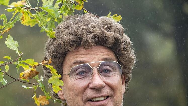 Ein Mann mit lockigem Haar und Brille steht im Freien und lächelt. Im Hintergrund sind grüne Blätter und Regentropfen zu sehen.