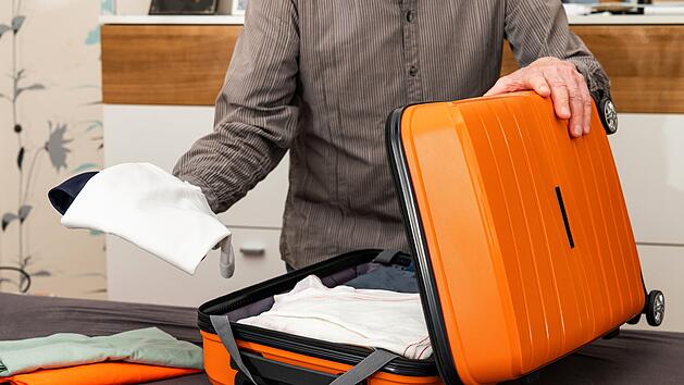 A man folds things in an orange-colored travel suitcase on wheels. Travel preparation.Ein Mann faltet Dinge in einem orangefarbenen Reisekoffer auf Rädern. Reisevorbereitung.