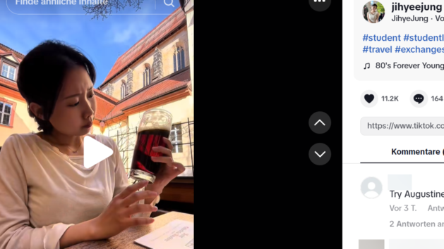 Eine Frau betrachtet verwundert ein Glas mit dunkler Fl&uuml;ssigkeit in einem sonnigen Hof. Im Hintergrund ist eine historische Geb&auml;udeansicht zu sehen.