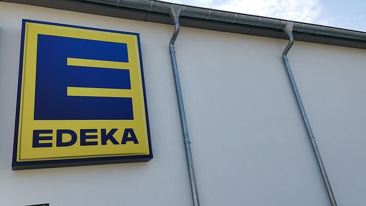Ein Edeka-Logo an einer Geb&auml;udewand, dar&uuml;ber blauer Himmel. Markante Dachrinnen rahmen das Bild ein.