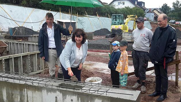Gut beschirmt von Architekt Rudolf R&ouml;big (links) nahm Mottens B&uuml;rgermeisterin Katja Habersack bei der Grundsteinlegung im Dauerregen die Maurerkelle selbst in die Hand. Interessierte Zuschauer waren Pfarrer Hans Thurn (rechts), Polier Florian Schad von der Baufirma Hodes sowie die beiden Buben Oskar Burth und Julian Darmer. Foto: Rolf Pralle