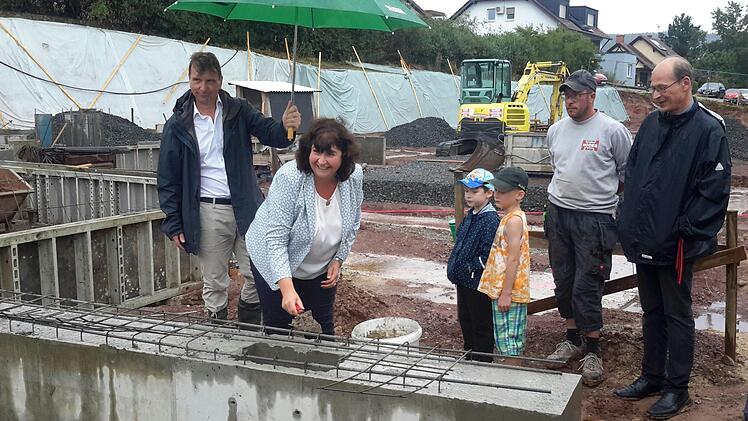 Gut beschirmt von Architekt Rudolf Röbig (links) nahm Mottens Bürgermeisterin Katja Habersack bei der Grundsteinlegung im Dauerregen die Maurerkelle selbst in die Hand. Interessierte Zuschauer waren Pfarrer Hans Thurn (rechts), Polier Florian Schad von der Baufirma Hodes sowie die beiden Buben Oskar Burth und Julian Darmer. Foto: Rolf Pralle