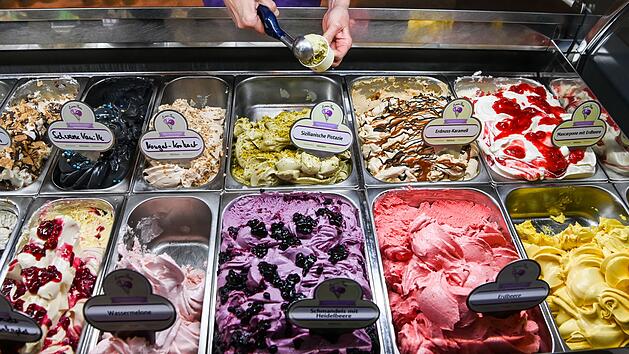 Eine Auswahl an Eissorten in einer Eisdiele, darunter Schwarze Vanille, Sizilianische Pistazie und Erdbeer. Eine Kugel Eis wird gerade serviert.