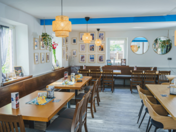 Gem&uuml;tliches Lokal mit Holztischen und St&uuml;hlen, dekoriert mit Bildern an wei&szlig;en W&auml;nden, modernen Lampen und Pflanzen. Ideal f&uuml;r gesellige Runden oder entspannte Mahlzeiten.