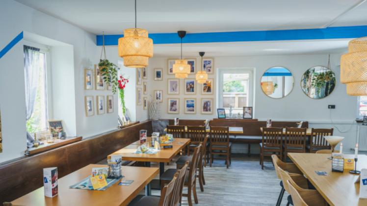 Gem&uuml;tliches Lokal mit Holztischen und St&uuml;hlen, dekoriert mit Bildern an wei&szlig;en W&auml;nden, modernen Lampen und Pflanzen. Ideal f&uuml;r gesellige Runden oder entspannte Mahlzeiten.