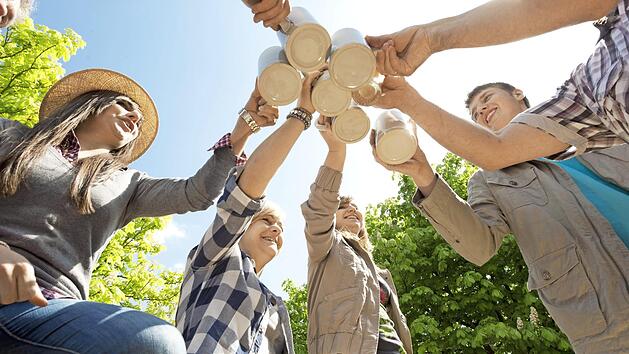 Menschen stoßen bei einem fröhlichen Outdoor-Treffen mit Bierkrügen an, umgeben von grünen Bäumen und blauem Himmel. Feierliche Stimmung, passend zum Brauereienlauf Nord 2025.