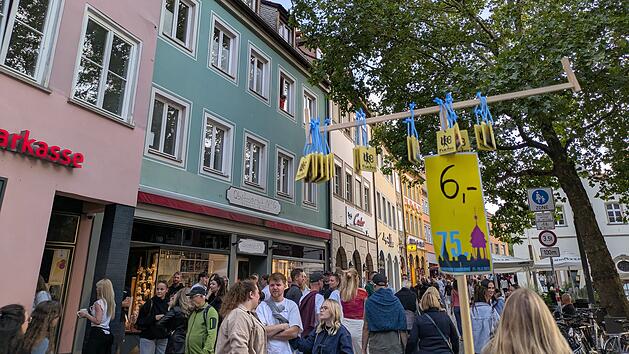 Eine Menschenmenge auf einem Festival, bunte H&auml;user im Hintergrund. Ein Schild weist auf einen 6-Euro-Festabzeichenpreis hin. B&auml;ume und Gesch&auml;fte s&auml;umen die Stra&szlig;e.