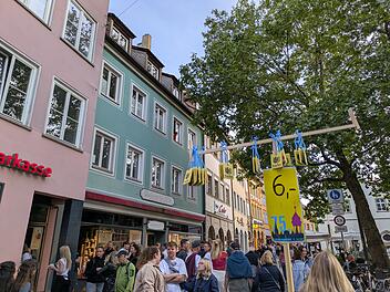 Eine Menschenmenge auf einem Festival, bunte Häuser im Hintergrund. Ein Schild weist auf einen 6-Euro-Festabzeichenpreis hin. Bäume und Geschäfte säumen die Straße.