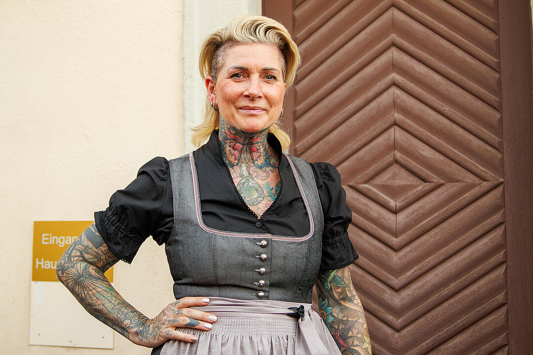 Eine t&auml;towierte Frau in einem Dirndl steht vor einer h&ouml;lzernen T&uuml;r. Sie l&auml;chelt leicht und hat eine hochgesteckte Frisur.