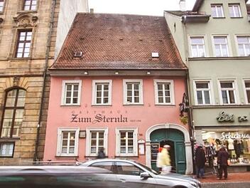 Das Bild zeigt das Gasthaus "Zum Sternla" in Bamberg, ein rosa Geb&auml;ude mit traditionellem Dach und Fenstern. Links und rechts schlie&szlig;en sich weitere historische H&auml;user an. Passanten sind zu sehen.