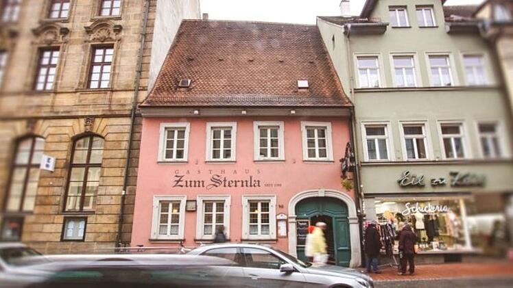 Das Bild zeigt das Gasthaus "Zum Sternla" in Bamberg, ein rosa Geb&auml;ude mit traditionellem Dach und Fenstern. Links und rechts schlie&szlig;en sich weitere historische H&auml;user an. Passanten sind zu sehen.