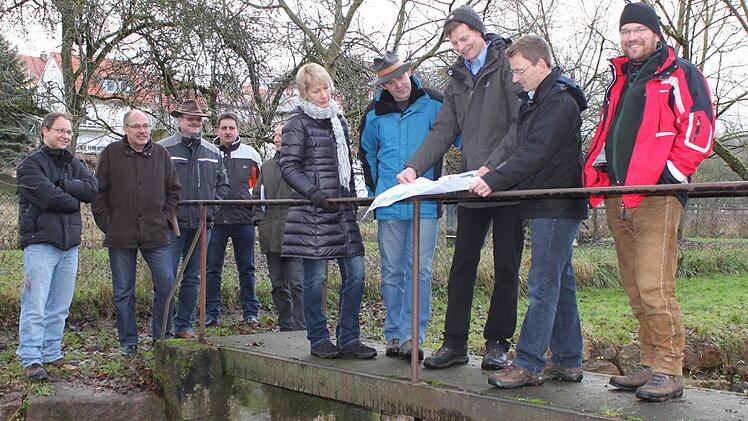 Martin Rottenberger (2. von rechts), Peter Borst (3. von rechts) und Wolf-Dieter Jacob (rechts) erklären den Praktikern aus den Gemeinden vor Ort ihre Pläne zur Einbeziehung des Nudelbachs in den Spielplatz in Hausen. Der soll in absehbarer Zeit saniert werden. Foto: Sabine Herteux