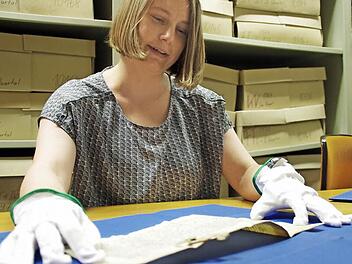 Über 600 Jahre alt ist dieses Pergament. Diese Frühmesse von 1381 ist eine der beiden ältesten Urkunden im Stadtarchiv. Das Schriftstück darf nur mit großer Vorsicht und Schutzhandschuhen berührt werden. Hinter Anja Weigelt türmen sich Kartons mit Zeitungen. Foto: Marco Meißner