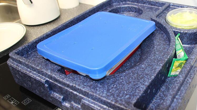 In dieser Styroporbox wird das Essen geliefert. Unter dem Porzellanteller mit blauem Deckel ist ein roter Akku, der die Mahlzeit warm hält. Foto: Dominic Buckreus
