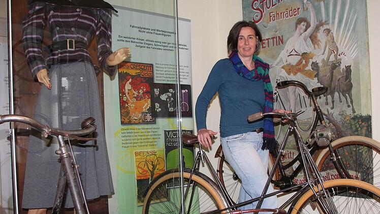 Frauen hinter dem Lenker: Links im Schaufenster steht eine Puppe mit so genannten Beinkleidern; links hängt ein Plakat, dass für Damenräder wirbt; in der Mitte steht Mona Buchmann vom Fahrradmuseum mit einem Modell für Frauen der Marke Pedersen, das sich besonders durch die filigrane Gitterkonstruktion auszeichnet.