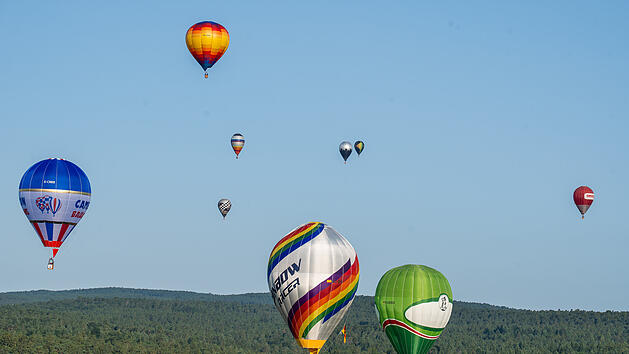 Mehrere bunte Heißluftballons schweben am blauen Himmel über einer bewaldeten Landschaft. Einige Ballons haben markante Muster und Logos. Die Szene wirkt friedlich und frei.