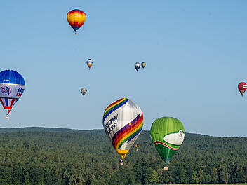 Mehrere bunte Heißluftballons schweben am blauen Himmel über einer bewaldeten Landschaft. Einige Ballons haben markante Muster und Logos. Die Szene wirkt friedlich und frei.